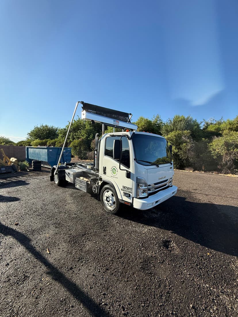 White roll-off truck with a crane arm parked in a gravel lot under clear blue sky.