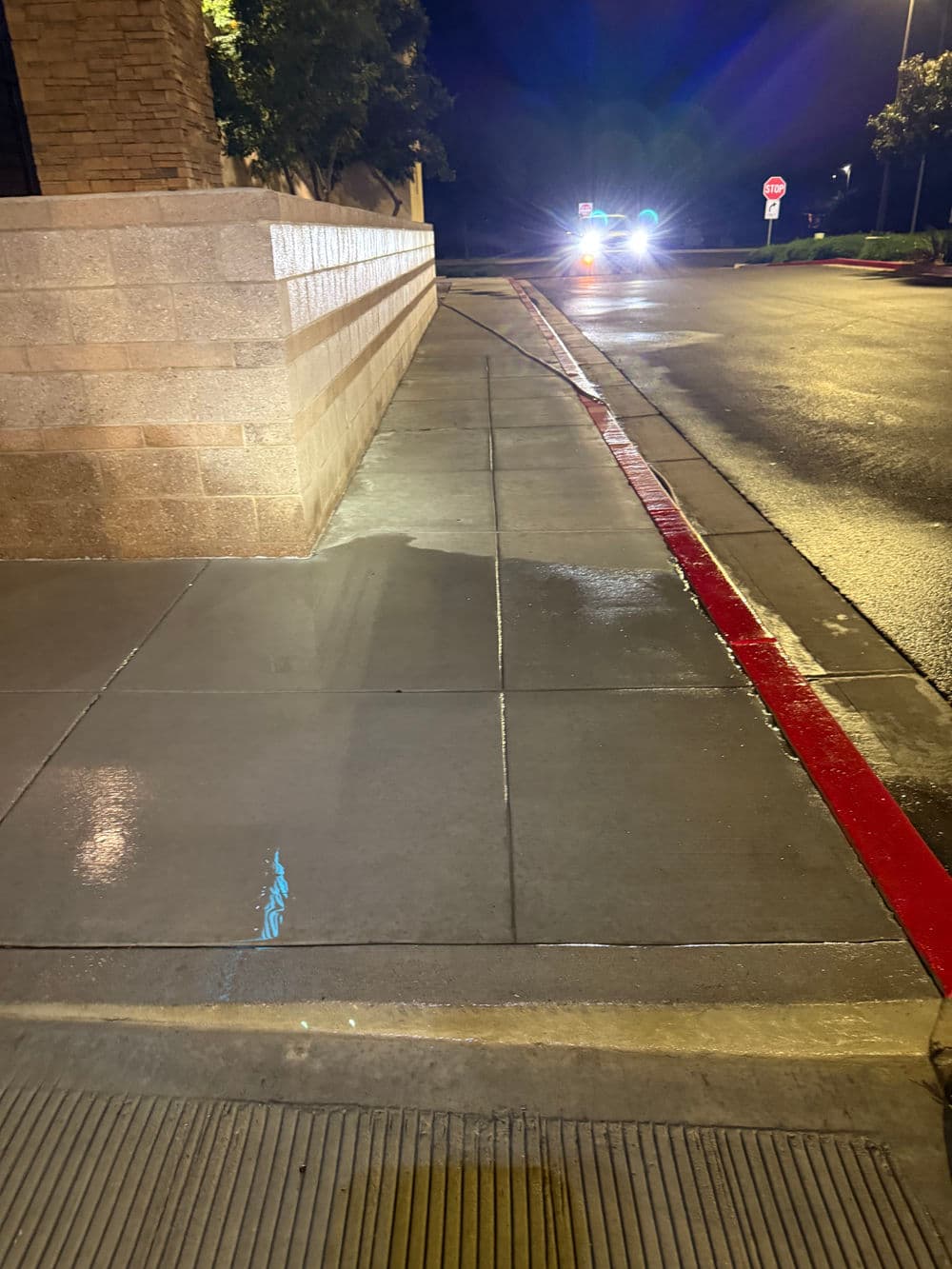 Wet sidewalk at night with a red curb and vehicle headlights in the distance.